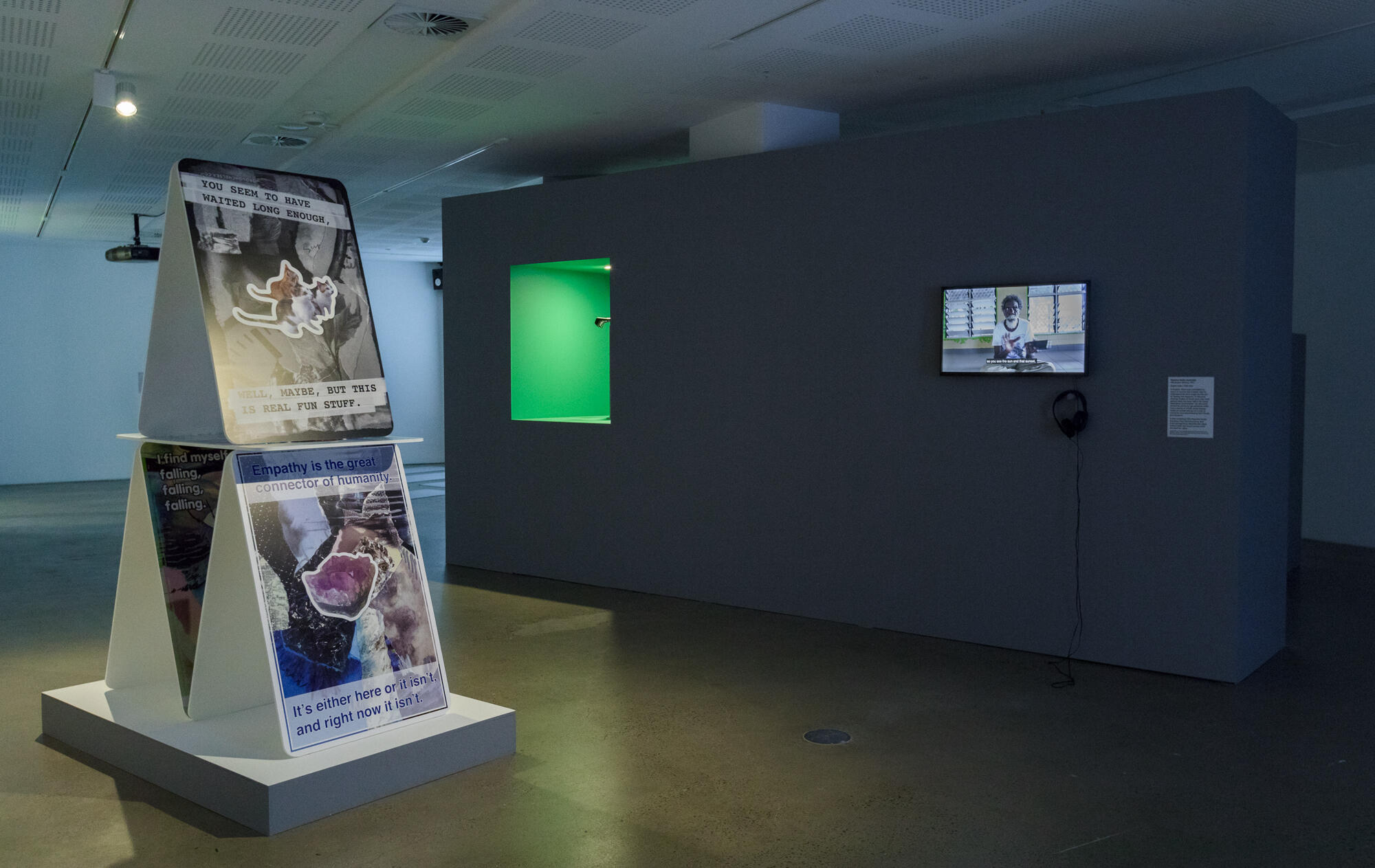 Installation view of a room containing a video, a green space built into a wall and very large cards stacked in the shape of a triangle.