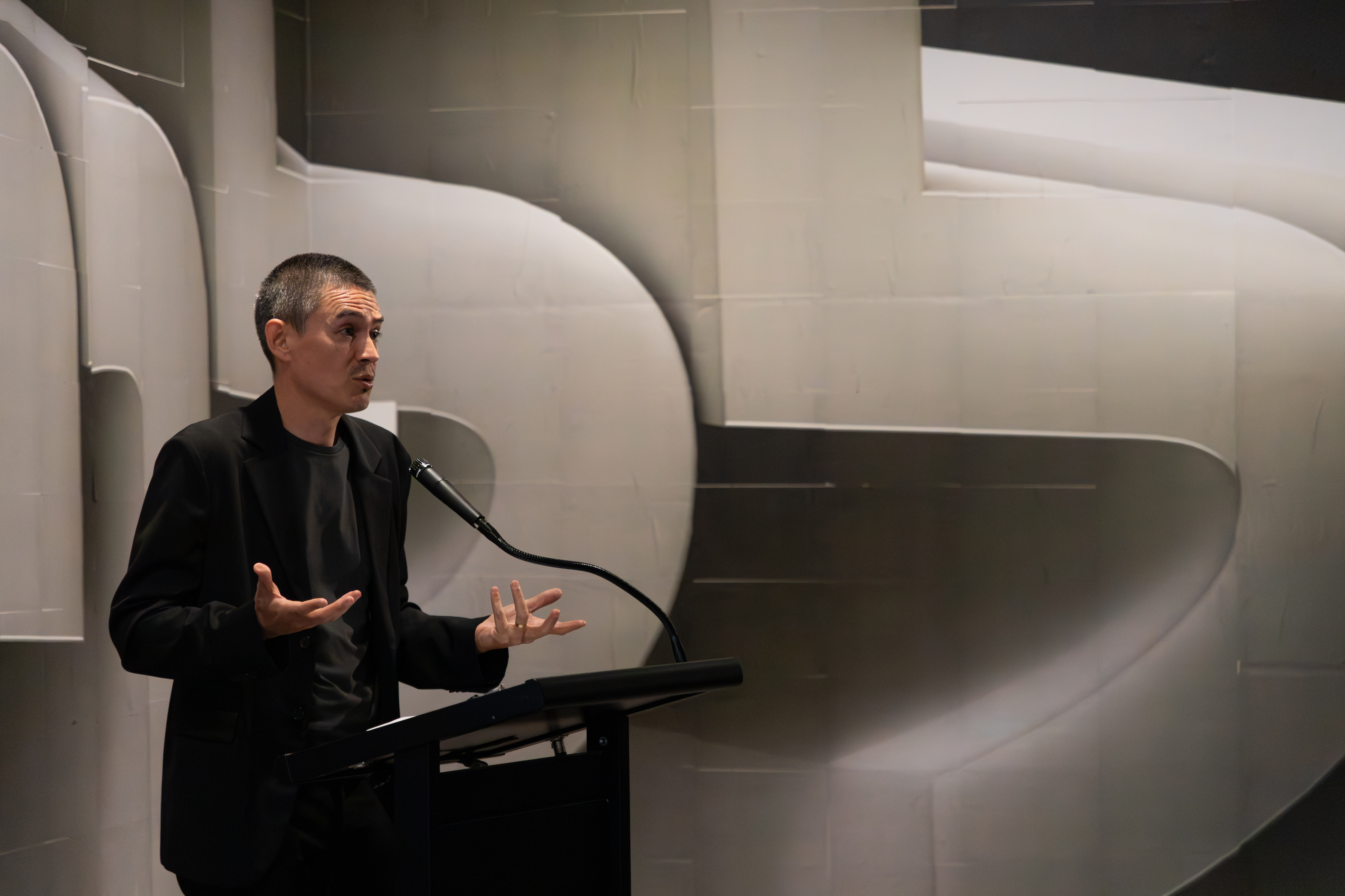 David Teh in a black outfit stands at a podium, gesturing while speaking.