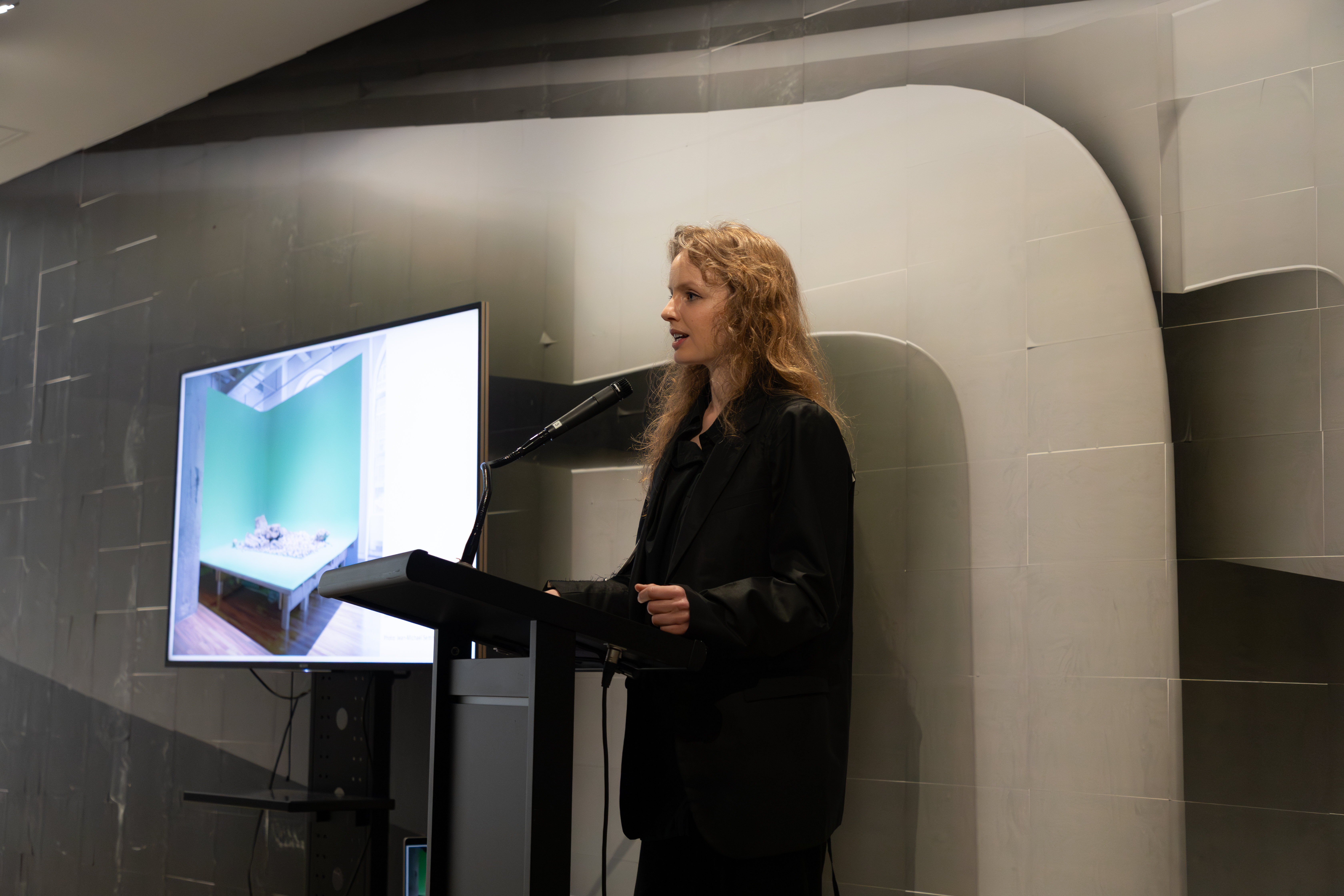  Stefanie Hessler stands at a podium, speaking into a microphone. They are dressed in a dark suit. A screen displays an image of a exhibition install.