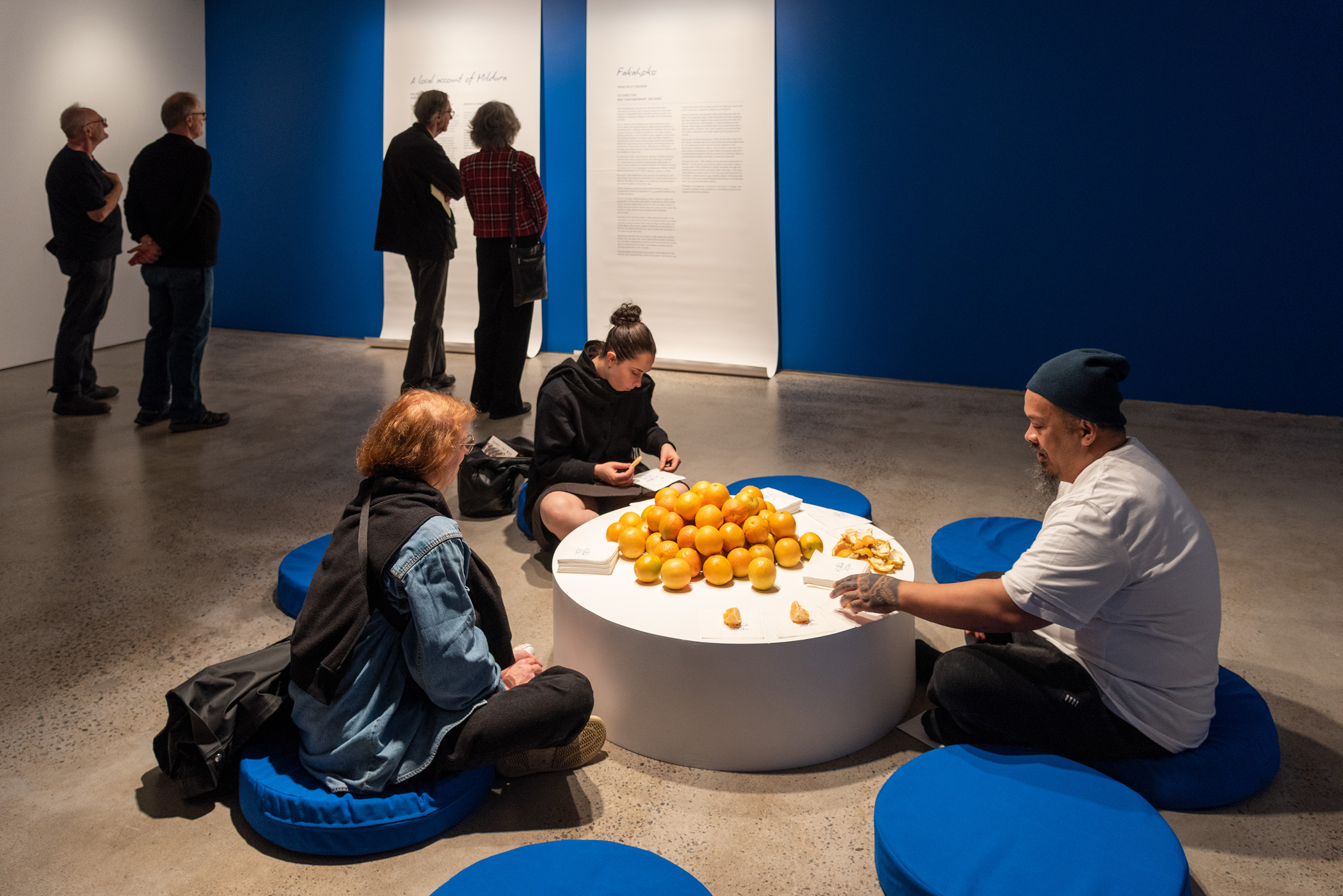 Kalisolaite ’Uhila with a pile of oranges and napkins, joined by two people.