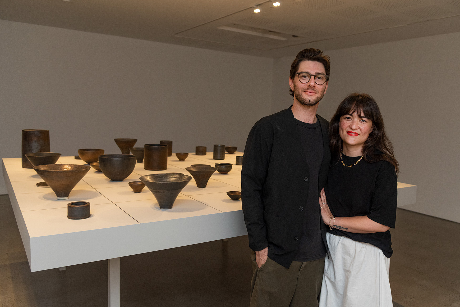 Artist, Jordan Gower and friend stand beside a large display of dark ceramic vessels arranged on a white table in a gallery.