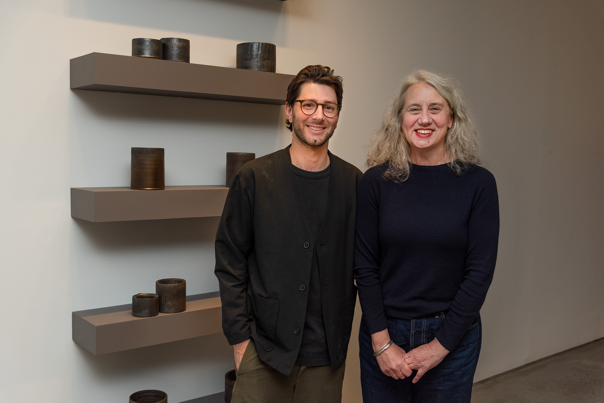 Artist, Jordan Gower and Kirsten Coelho stand beside a large display of dark ceramic vessels arranged on a white table in a gallery.