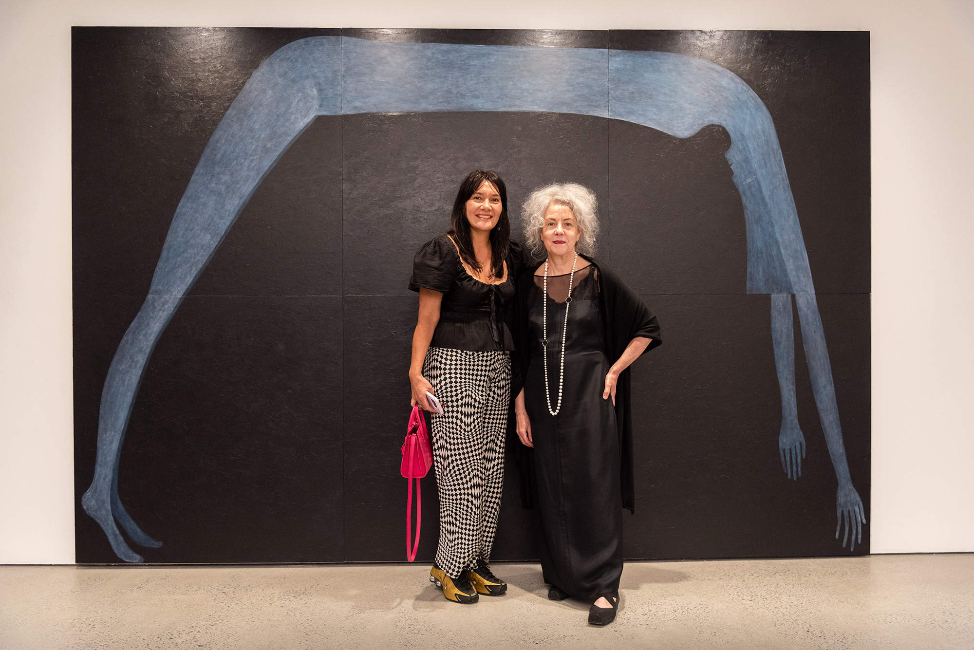 Artist Heather B. Swann and friend stand in front of a large multi-panel painting featuring an elongated blue figure against a black background.