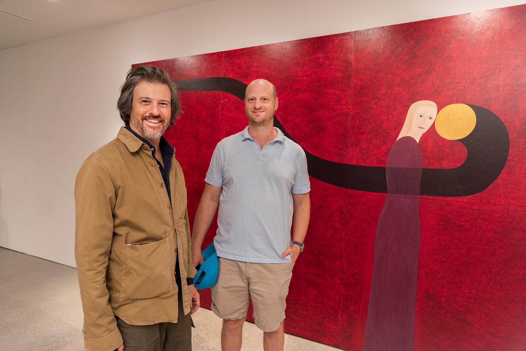 stand in front of a large pink painting depicting a pale figure beside a white circular table, posing within a gallery setting.
