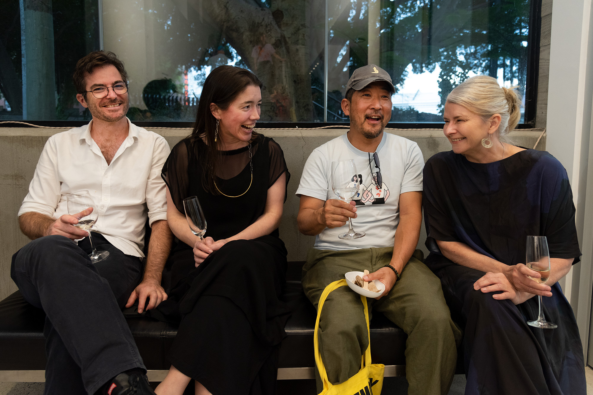 Four people sit together on a bench holding wine glasses and talking beneath a large window during a gallery opening.