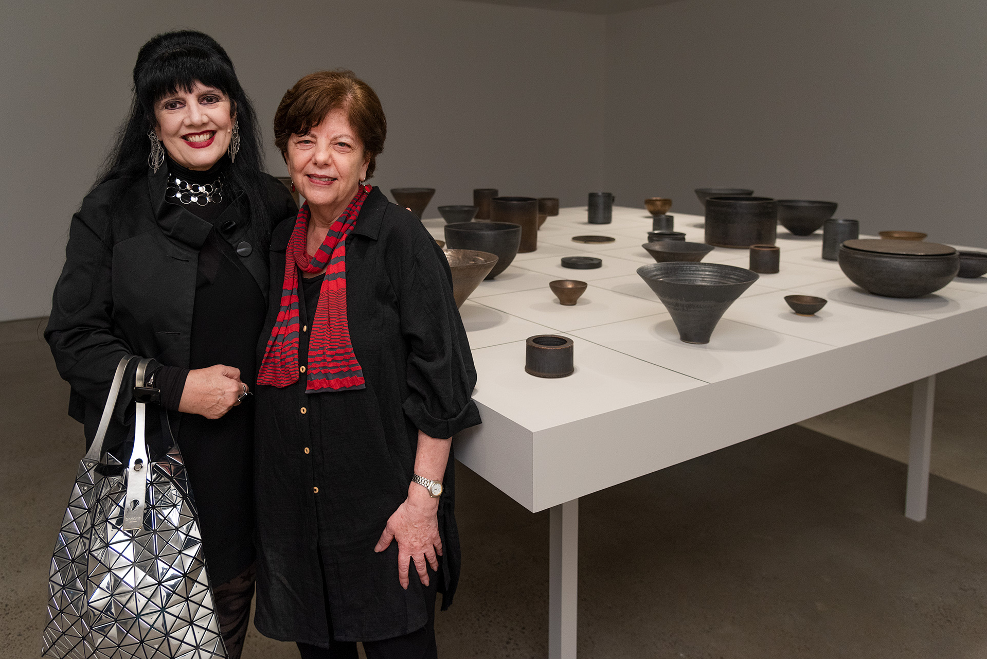 Two people stand beside a large white display table presenting a collection of dark ceramic vessels in a contemporary gallery space.