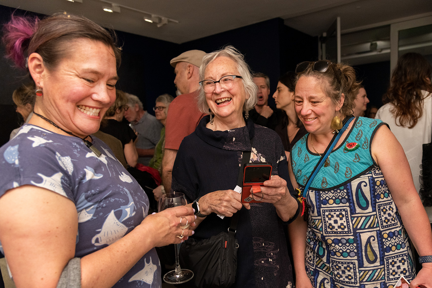 Three attendees converse and hold drinks while standing among a crowd at an exhibition opening in a gallery foyer.