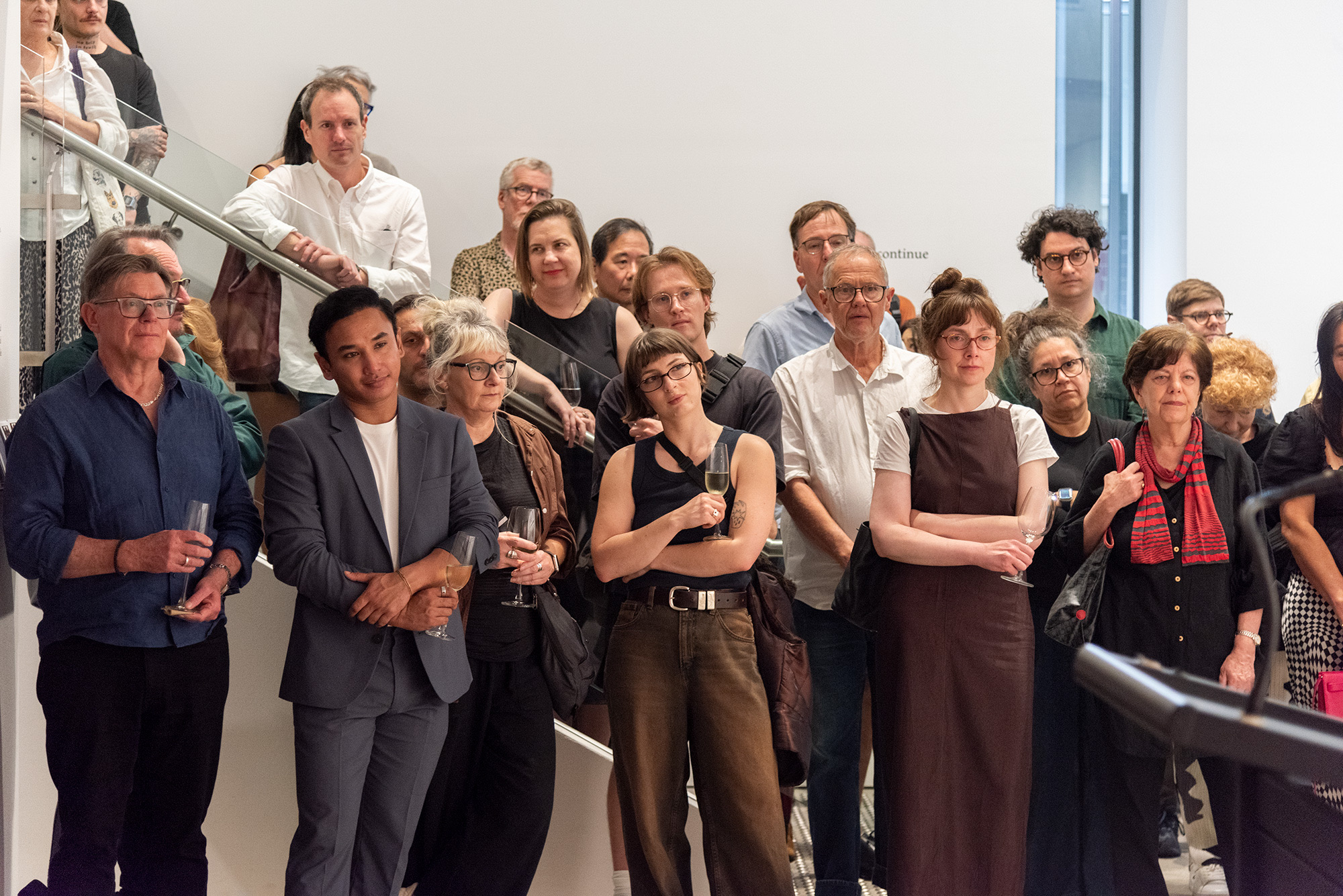 Visitors stand closely together listening to a speech during a gallery opening, many holding drinks and facing toward the speaker.