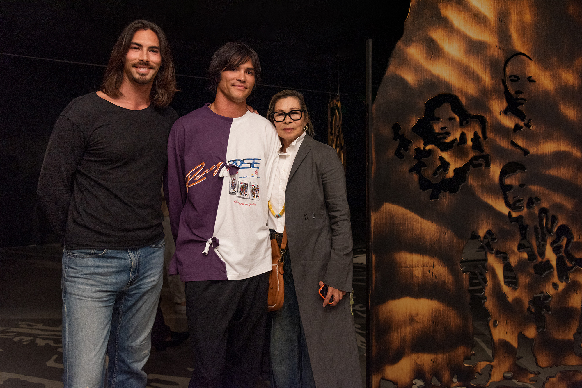 Artist Remy Faint and two people pose beside a large wooden panel with cut-out figures in a dimly lit gallery space.