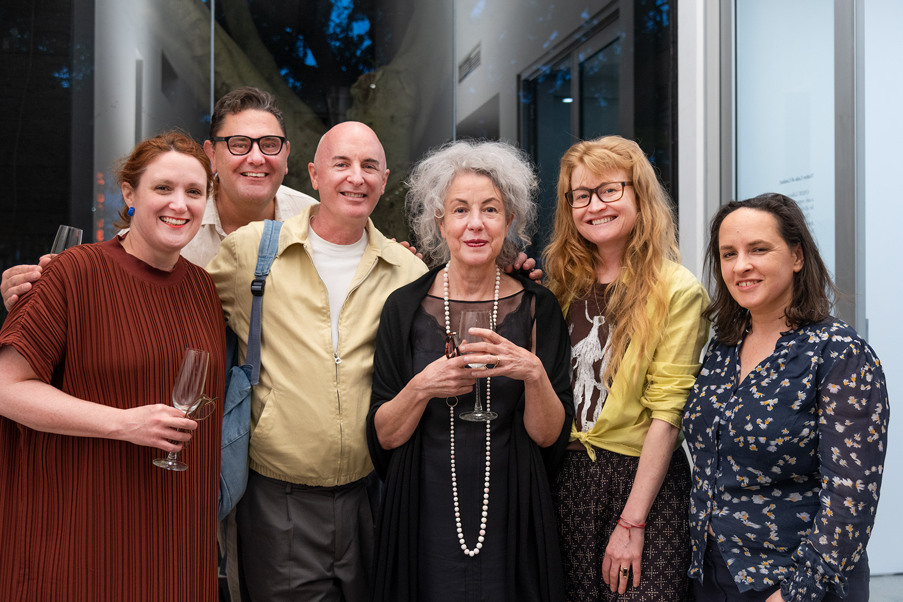 Artist Heather B. Swann and five guests pose together at a gallery reception, standing closely in front of large windows while two hold wine glasses.