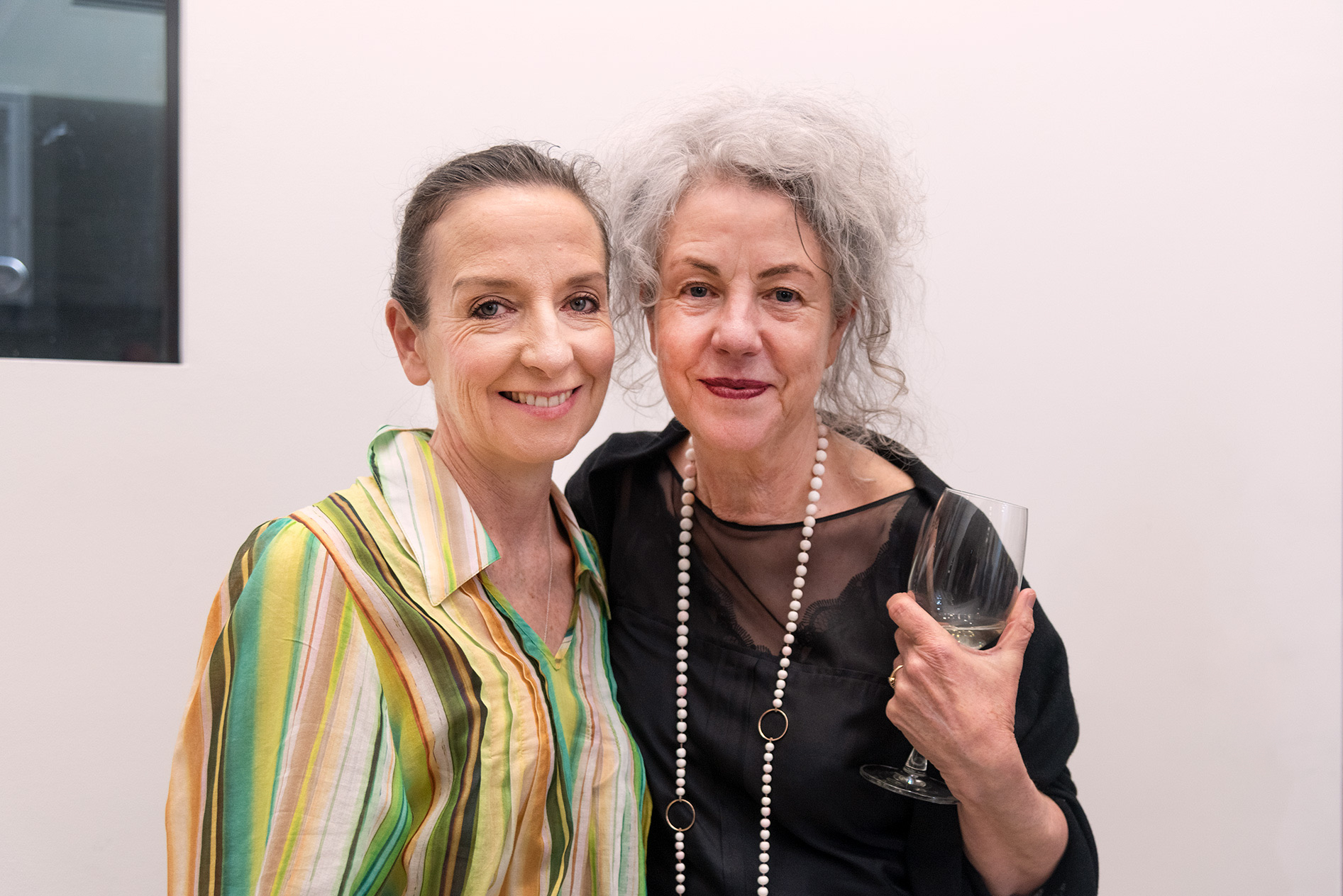 Artist Heather B. Swann adn friend stand side by side posing for a photograph at a gallery event, with one holding an empty wine glass against a plain white gallery wall.
