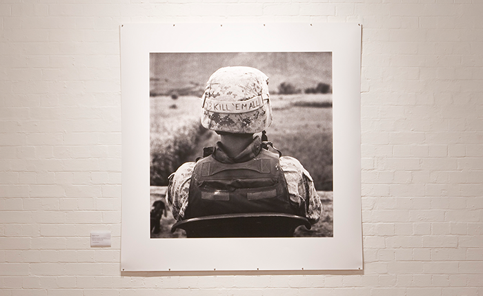 Black-and-white documentary-style photograph mounted as a gallery print on a white brick wall, showing the back view of a soldier seated outdoors.