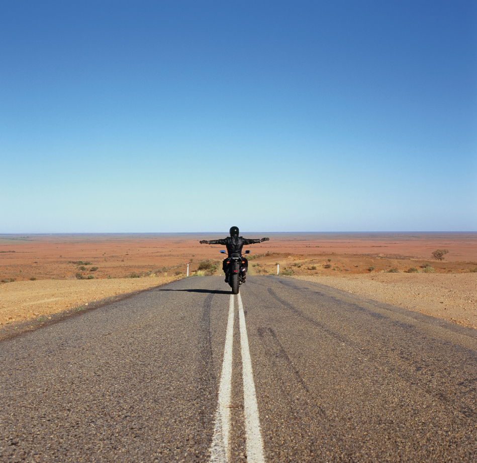 Still of a video work. Motorcyclist riding down a straight empty desert road with arms outstretched, disappearing into the horizon under a clear blue sky.