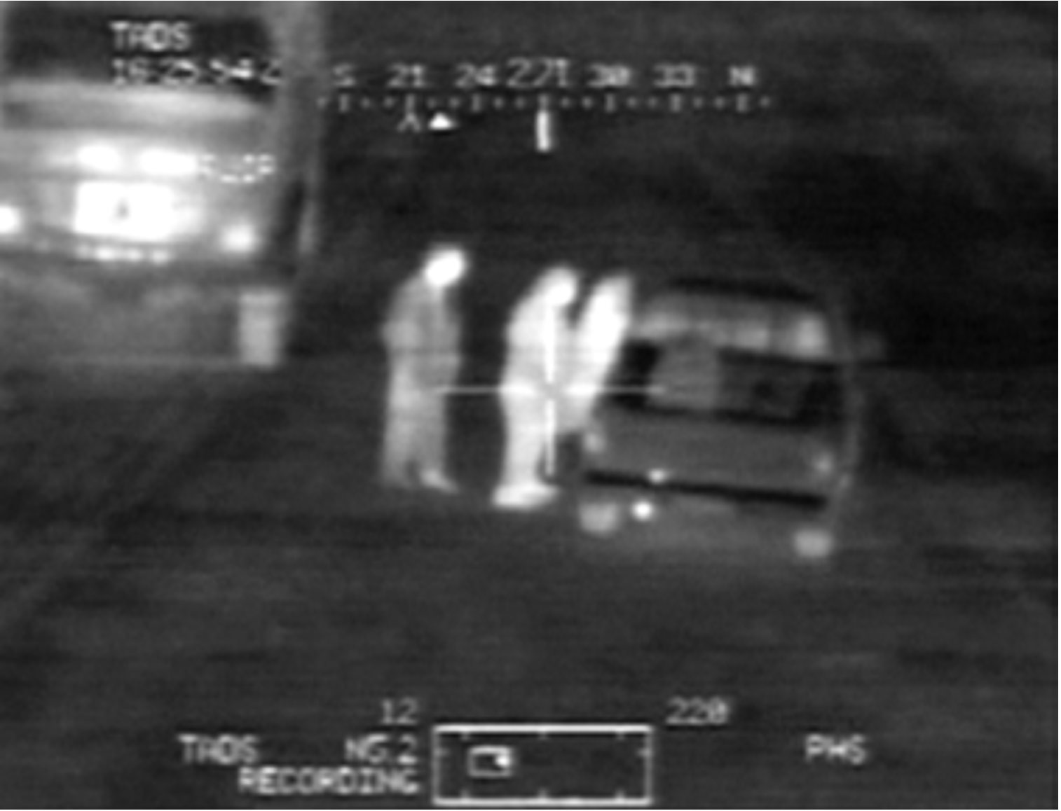 Still from video work displaying grainy black-and-white thermal/night-vision surveillance showing a roadside scene at night, with three human figures rendered as bright white heat signatures standing beside a parked vehicle on the right while another vehicle with intense headlights is visible on the left.