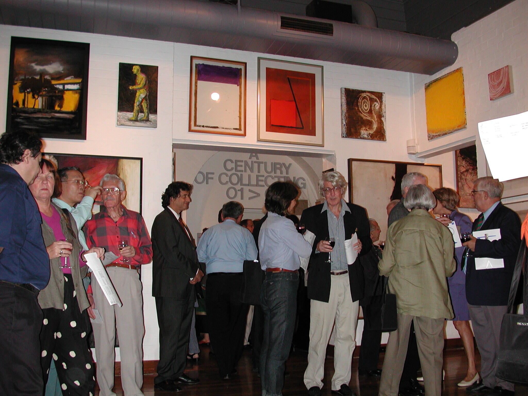 Wide view of a contemporary art gallery exhibition featuring abstract paintings mounted on white brick walls, polished wooden floors, and an open reception area in the background.