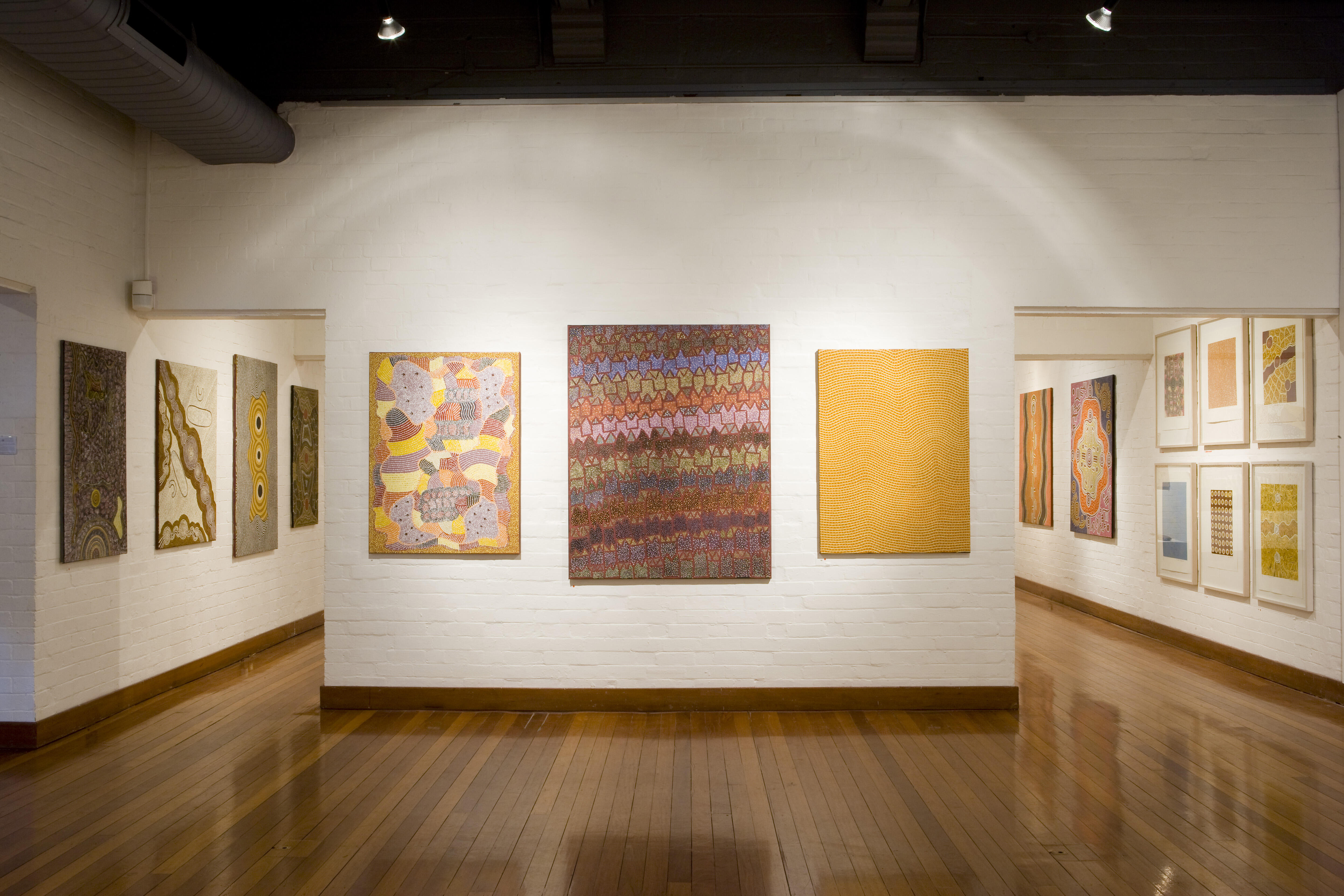 Installation view featuring contemporary Indigenous paintings displayed on white walls in a modern gallery space.