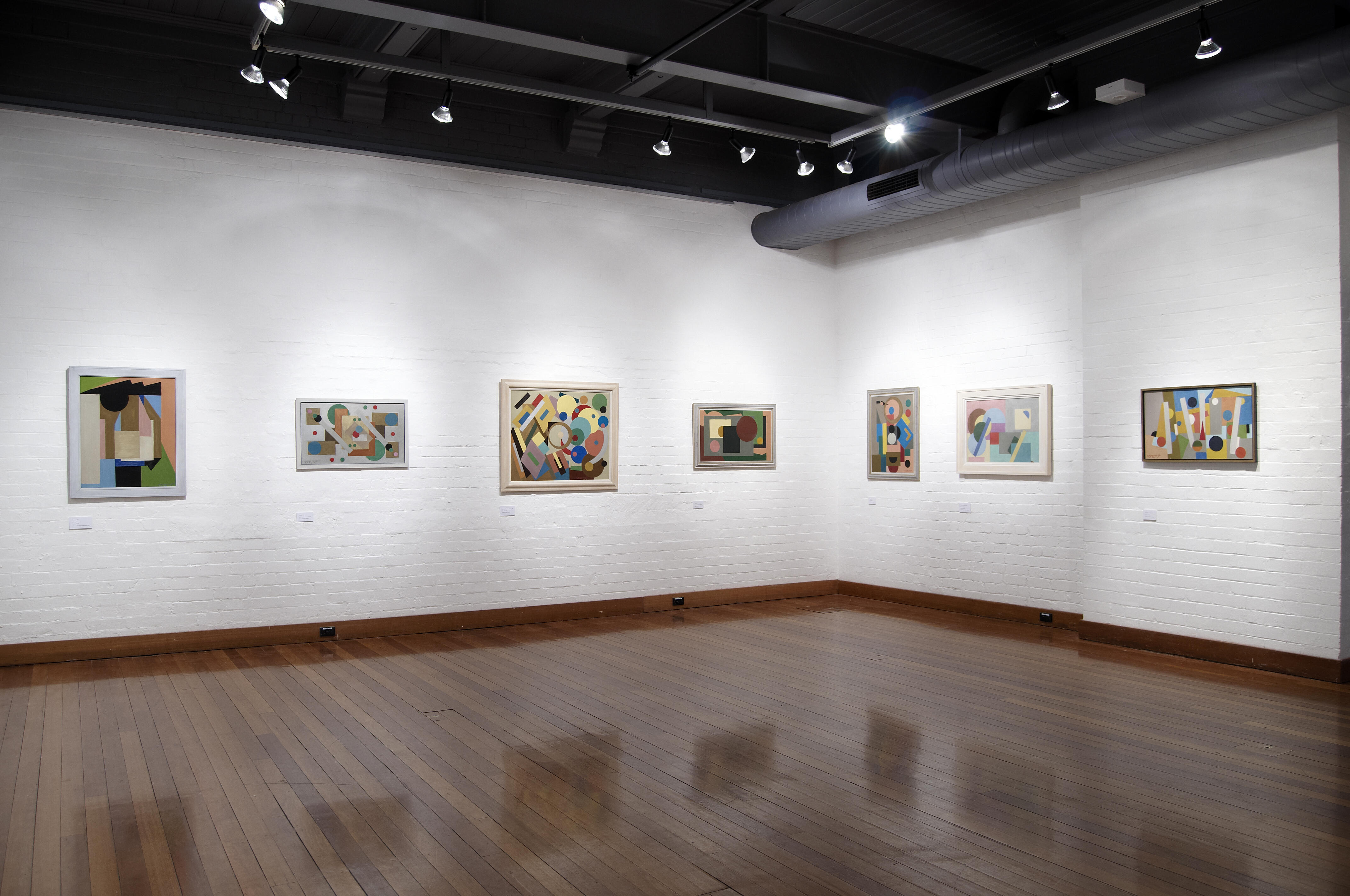 Display of framed abstract geometric paintings in black frames. The works feature bold circles, lines, and color blocks in red, blue, yellow, green, and brown tones.