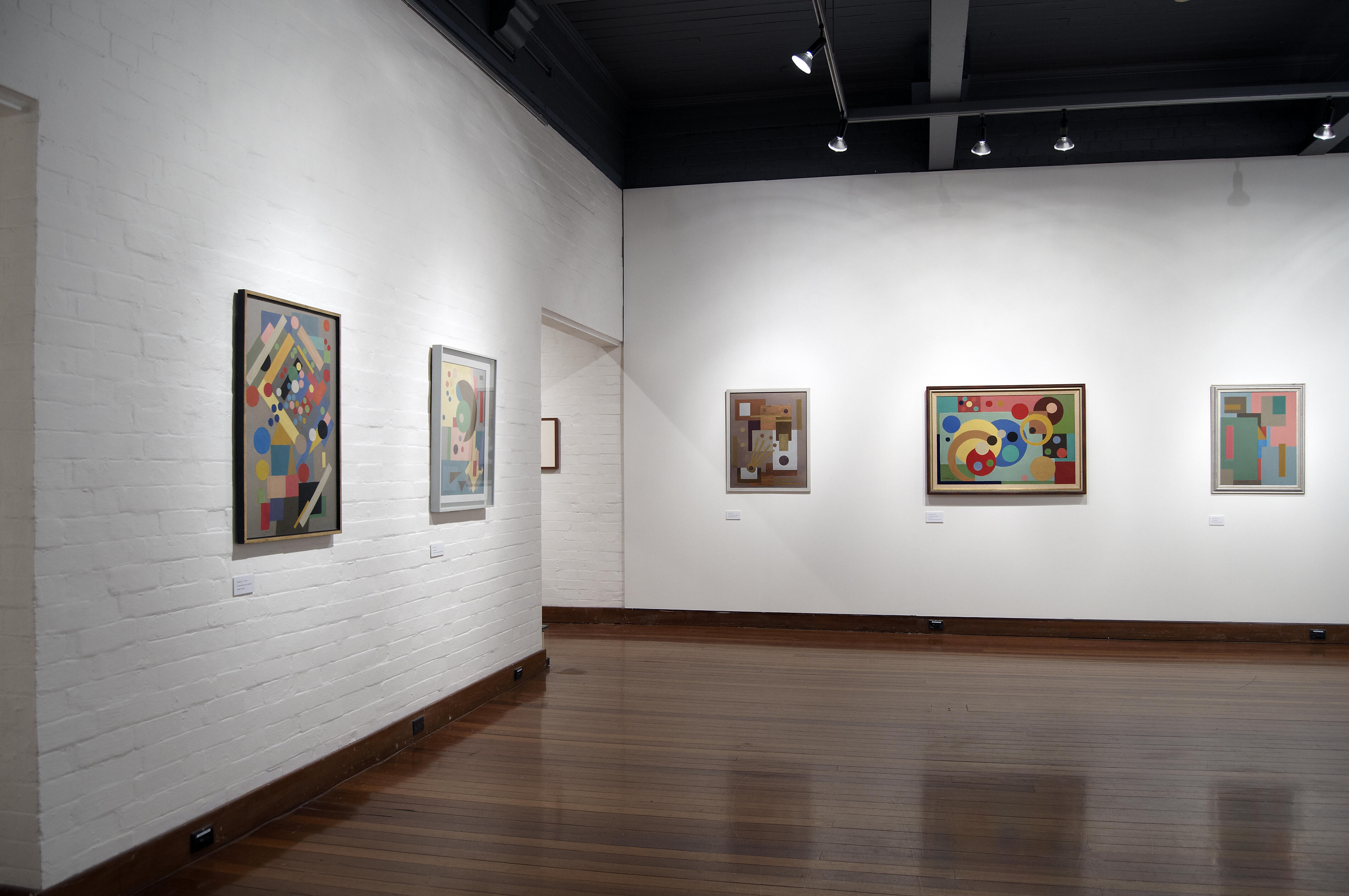 Framed abstract geometric paintings in black frames. The works feature bold circles, lines, and color blocks in red, blue, yellow, green, and brown tones.