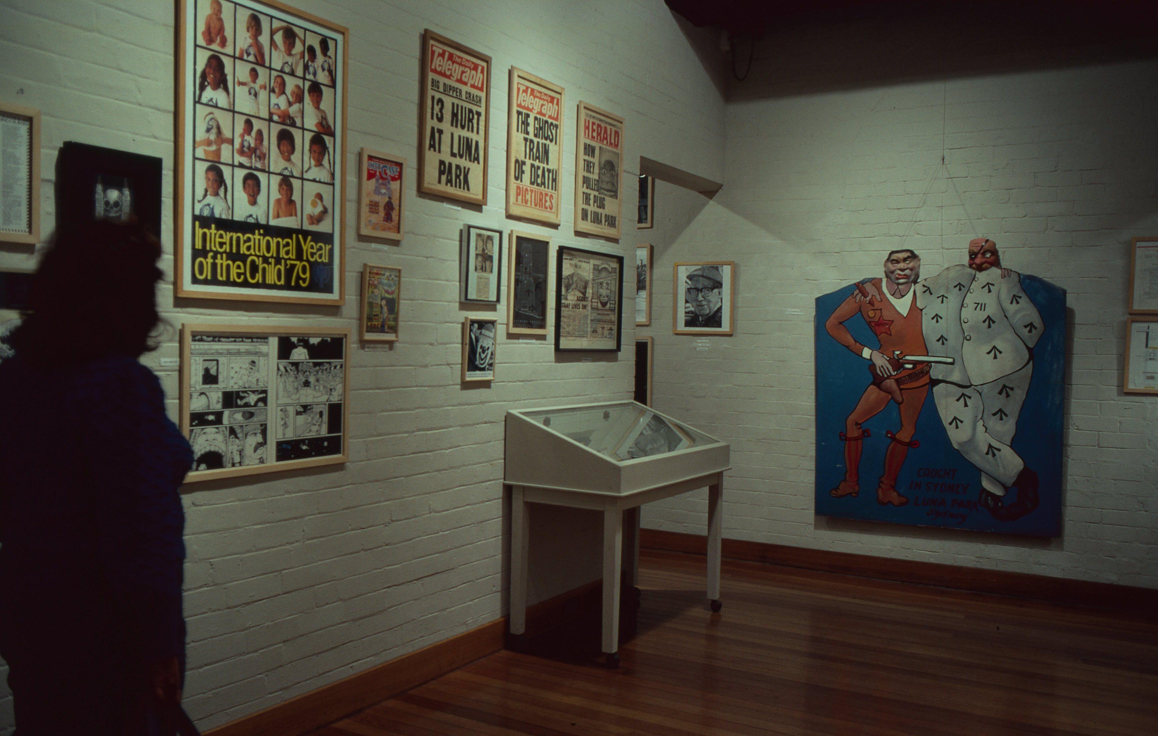Museum exhibit wall displaying framed posters, newspaper front pages, photographs, and comic artwork, with a glass display case in the center and a painted carnival-style figure hanging on the right wall. A visitor appears in silhouette at the left edge of the room.