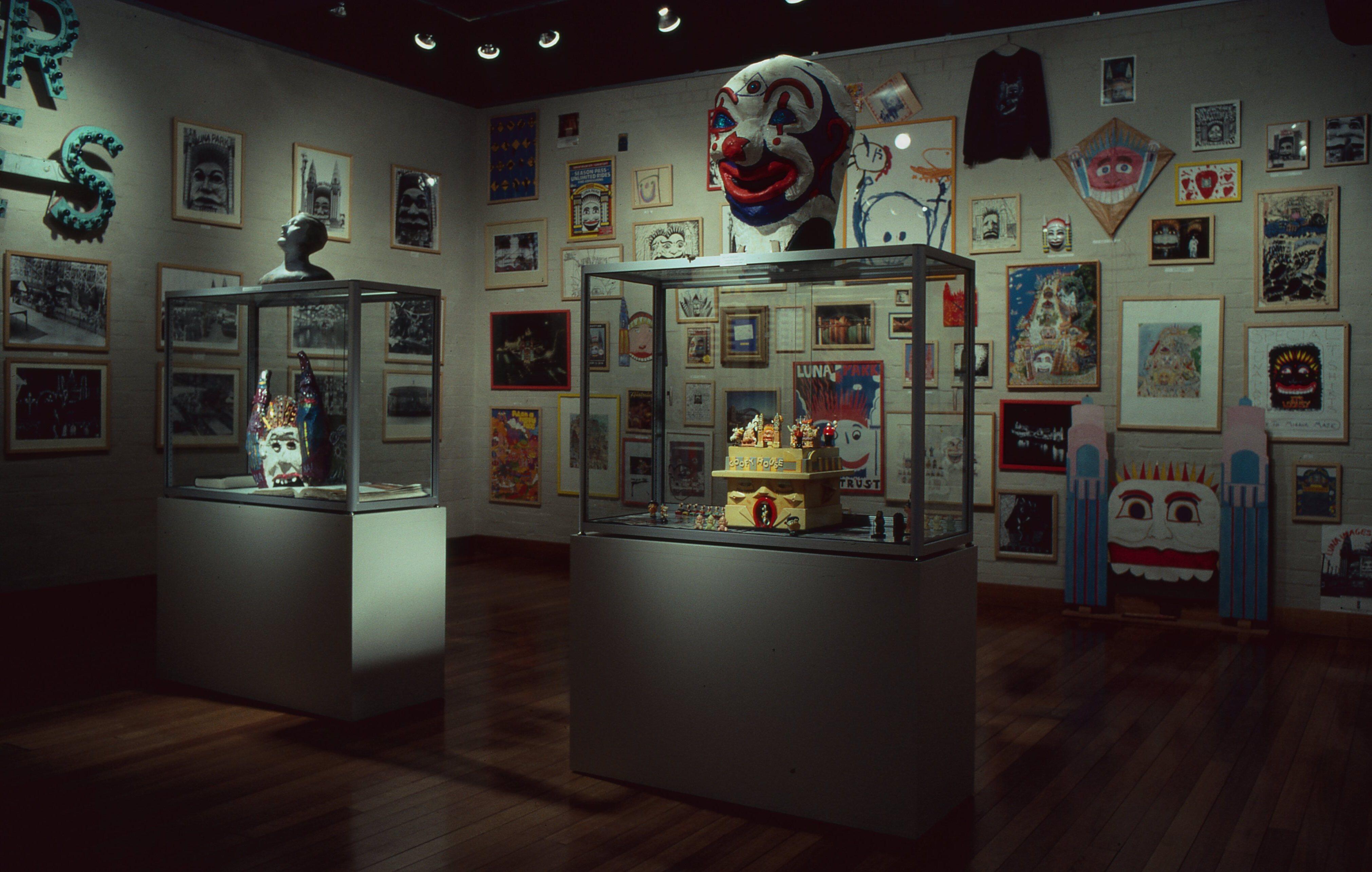 Dimly lit museum gallery featuring large cartoon-style murals on the walls, two glass display cases with artifacts in the center, and silhouettes of vintage automobiles mounted near the ceiling.