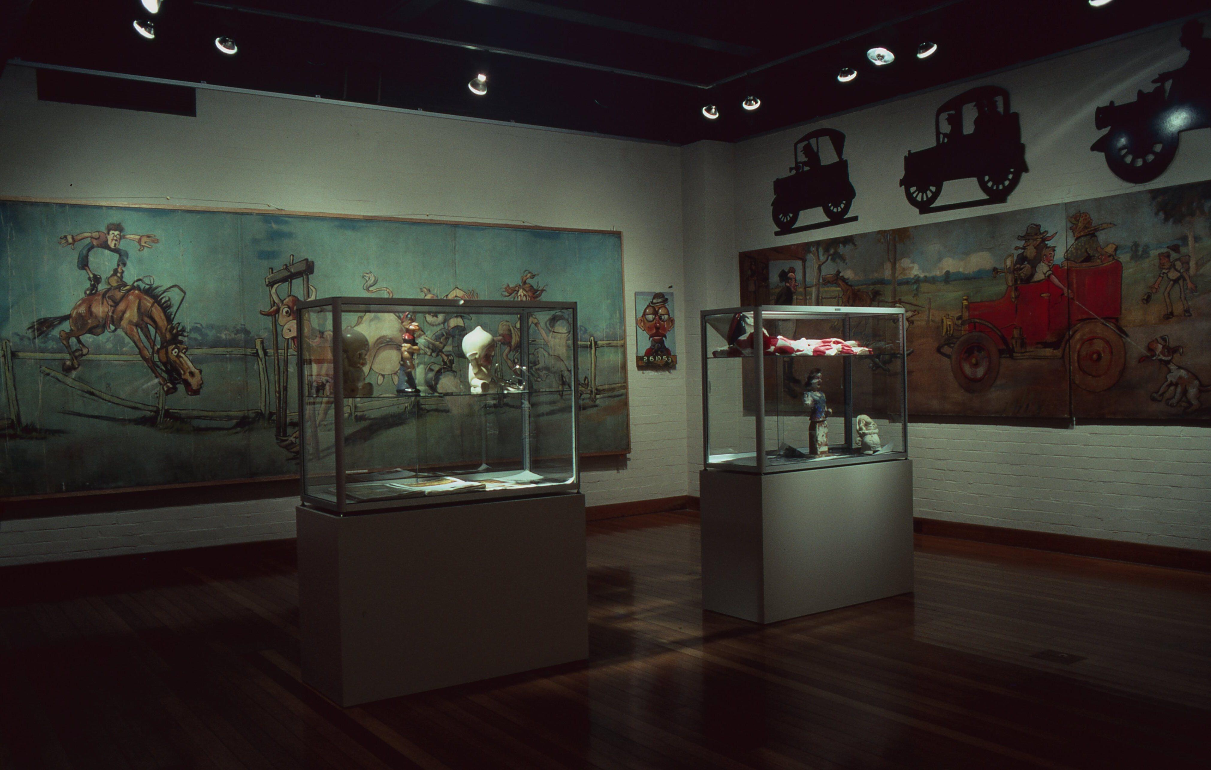 Dimly lit museum gallery featuring large cartoon-style murals on the walls, two glass display cases with artifacts in the center, and silhouettes of vintage automobiles mounted near the ceiling.