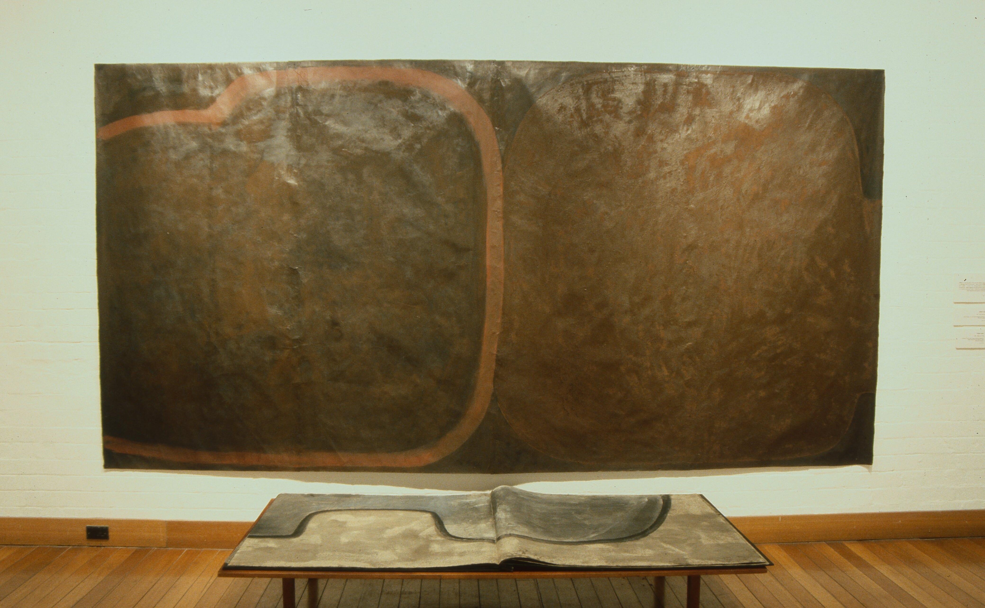 Large abstract artwork displayed in a gallery, featuring two rounded rectangular forms in dark brown and black tones with a muted reddish outline, mounted above a sculptural book-like piece on a wooden table.