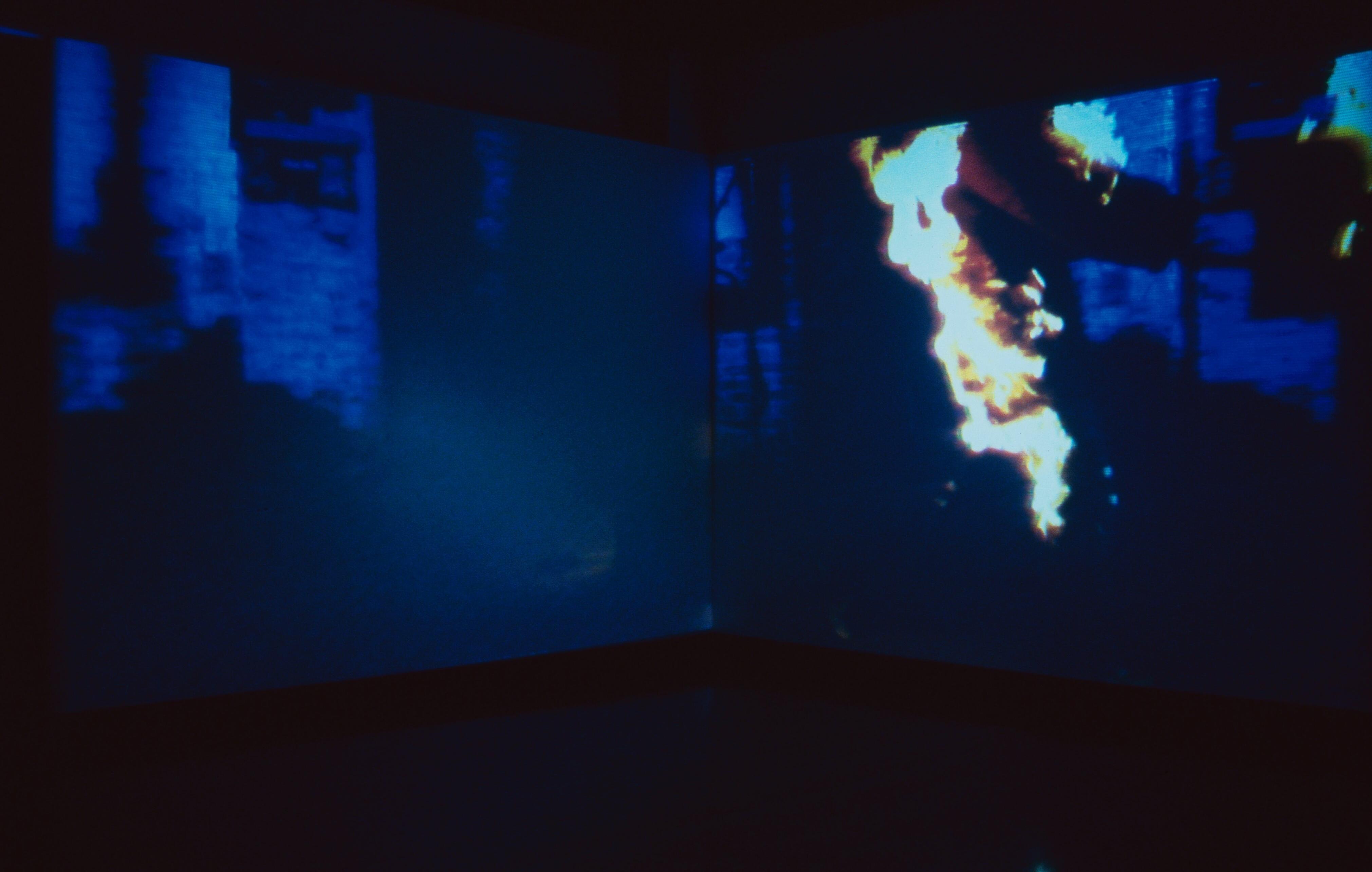Blue-toned video installation projected across two walls, showing a blurred figure in motion inside an industrial space.
