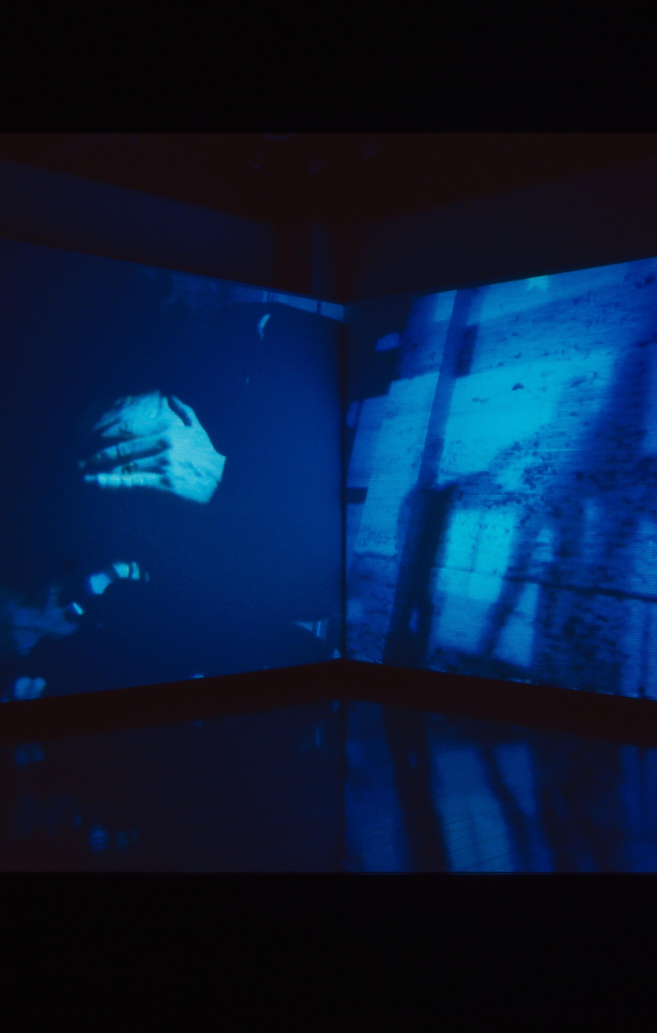 Blue-toned video installation projected across two walls, showing a blurred figure in motion inside an industrial space.