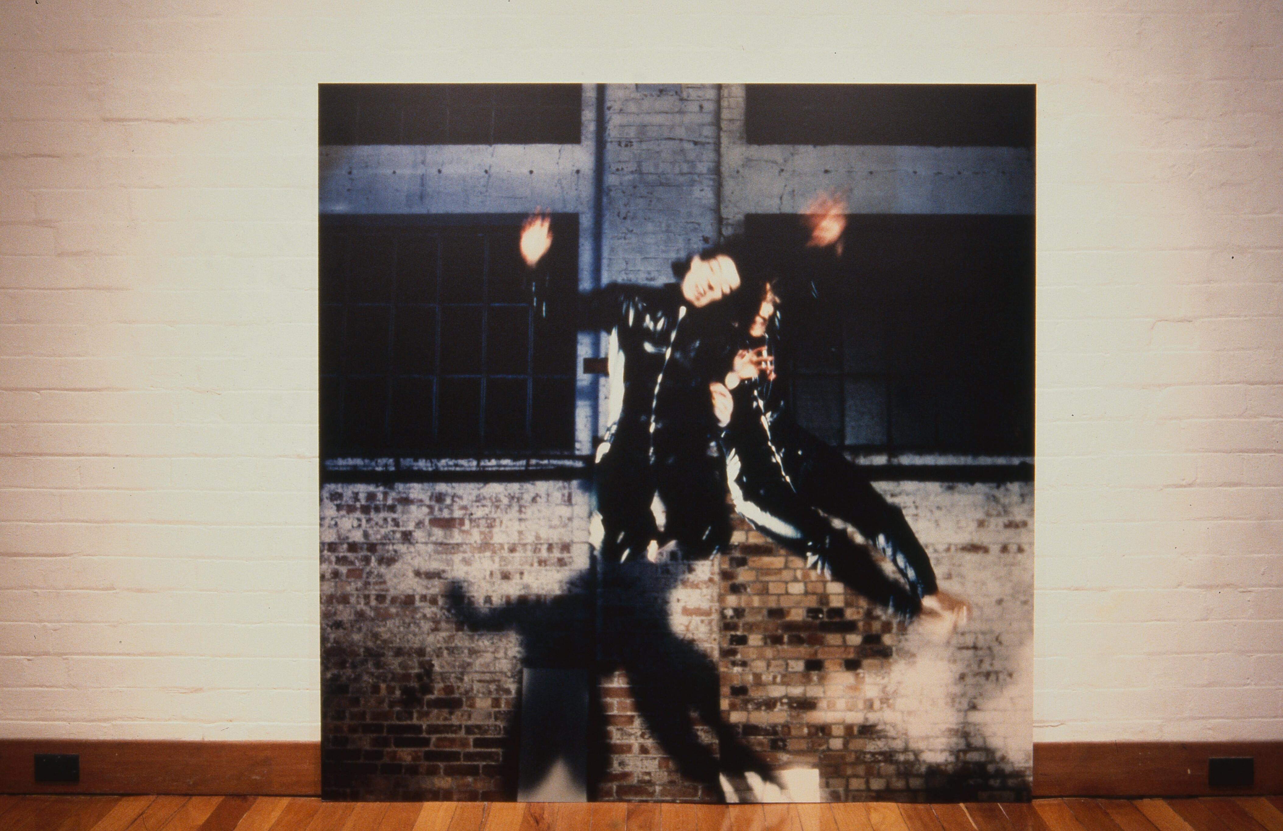 Photograph of two people in black clothing jumping in front of a brick wall with large windows, their motion blurred.