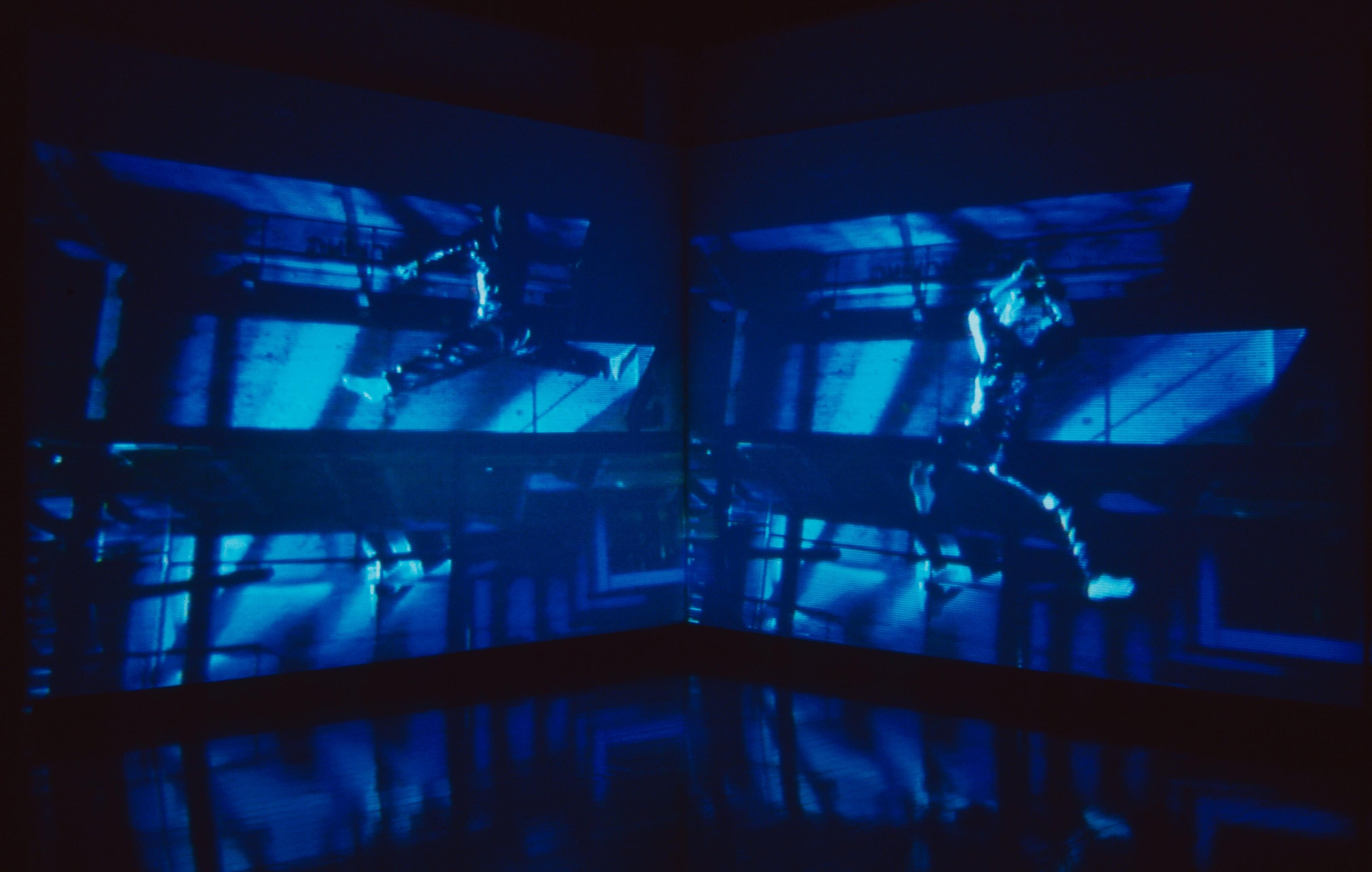 Blue-toned video installation projected across two walls, showing a blurred figure in motion inside an industrial space.