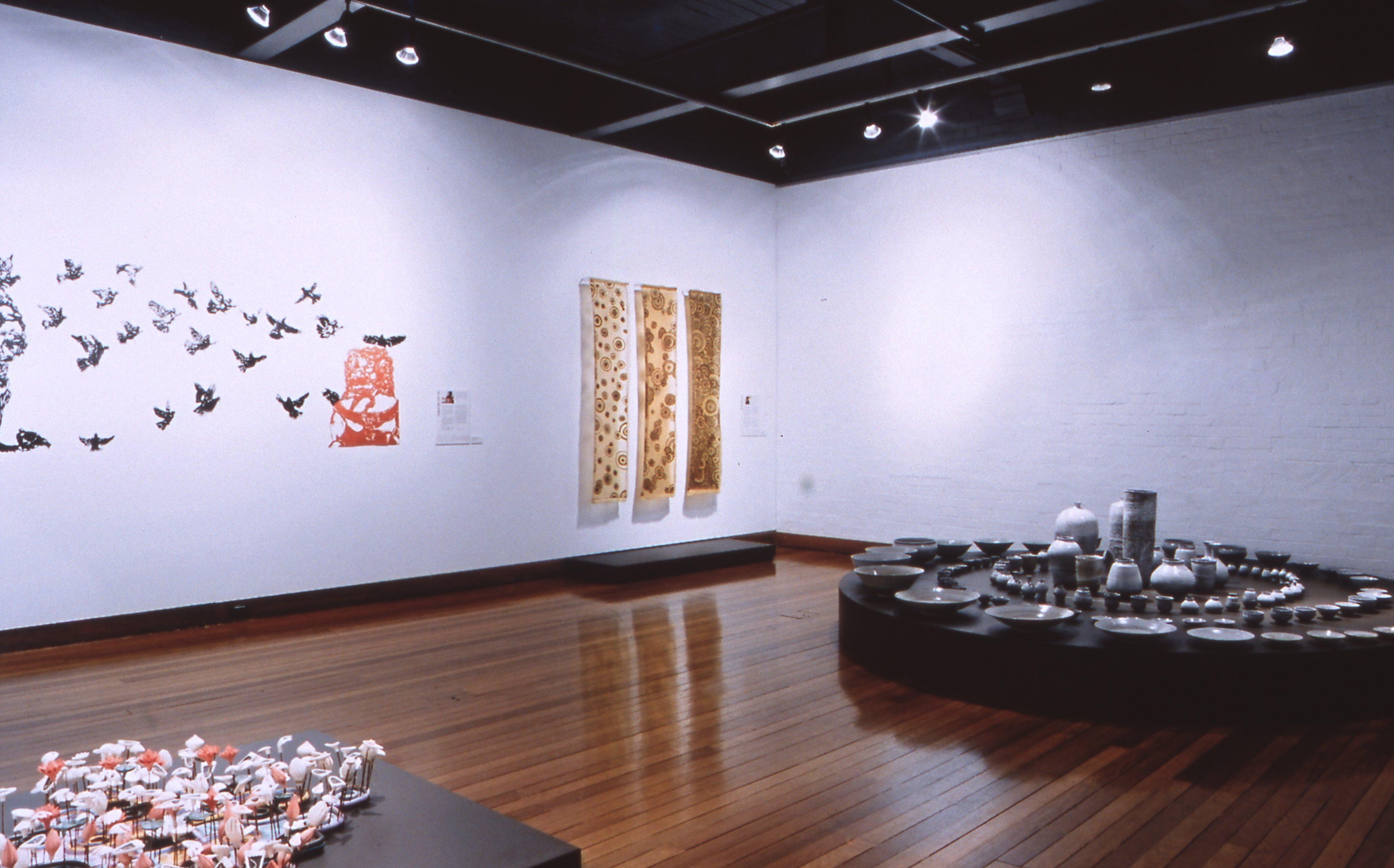 The composition features multiple sculptural and mixed-media works arranged across the floor and walls, including framed textile-like pieces, suspended or wall-mounted objects, and ceramic or organic forms.