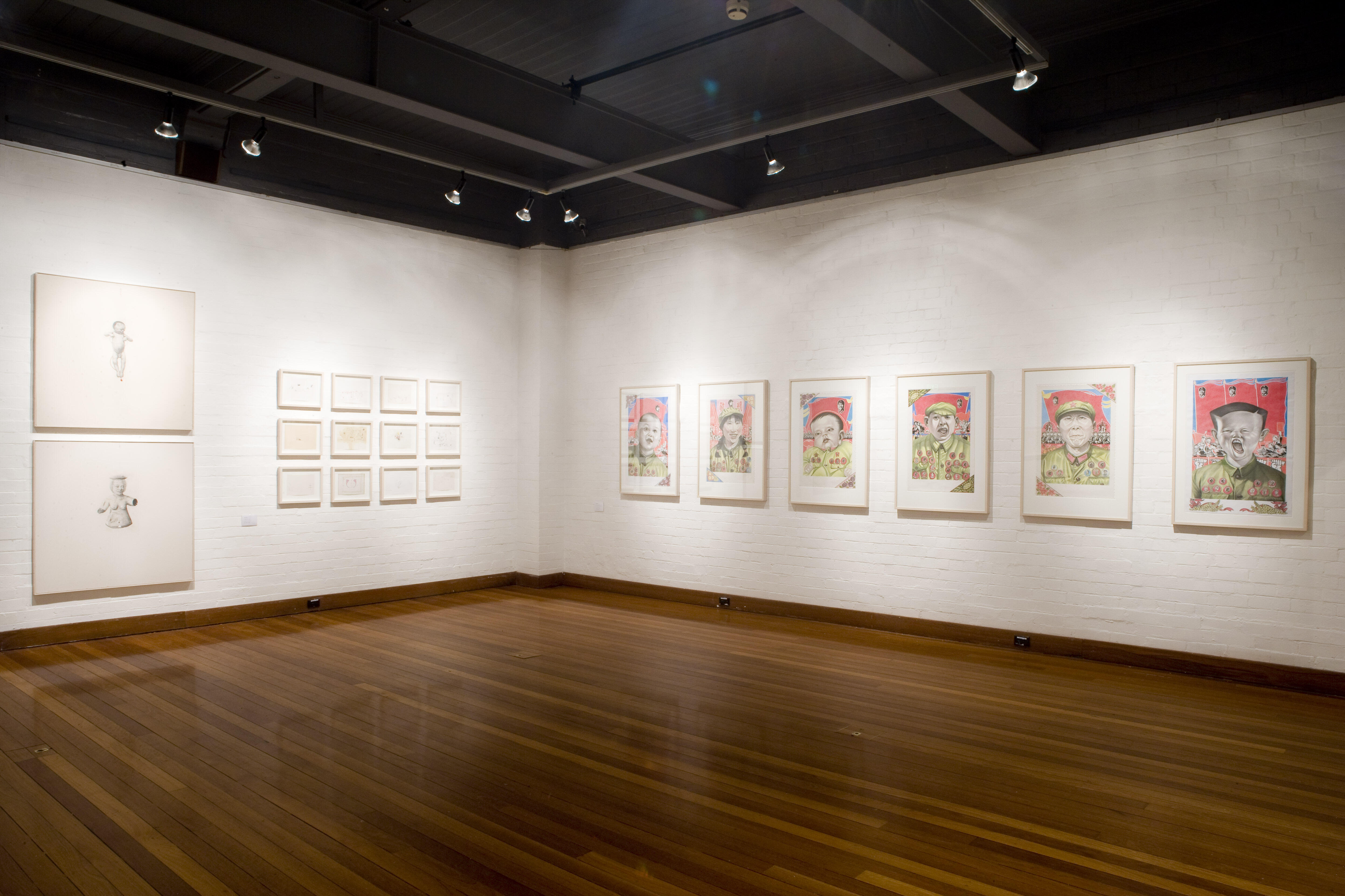 Interior with white brick walls, wooden floors, and track lighting, displaying framed series of colorful portrait prints on the right and monochrome sketches and drawings arranged in grids on the left.