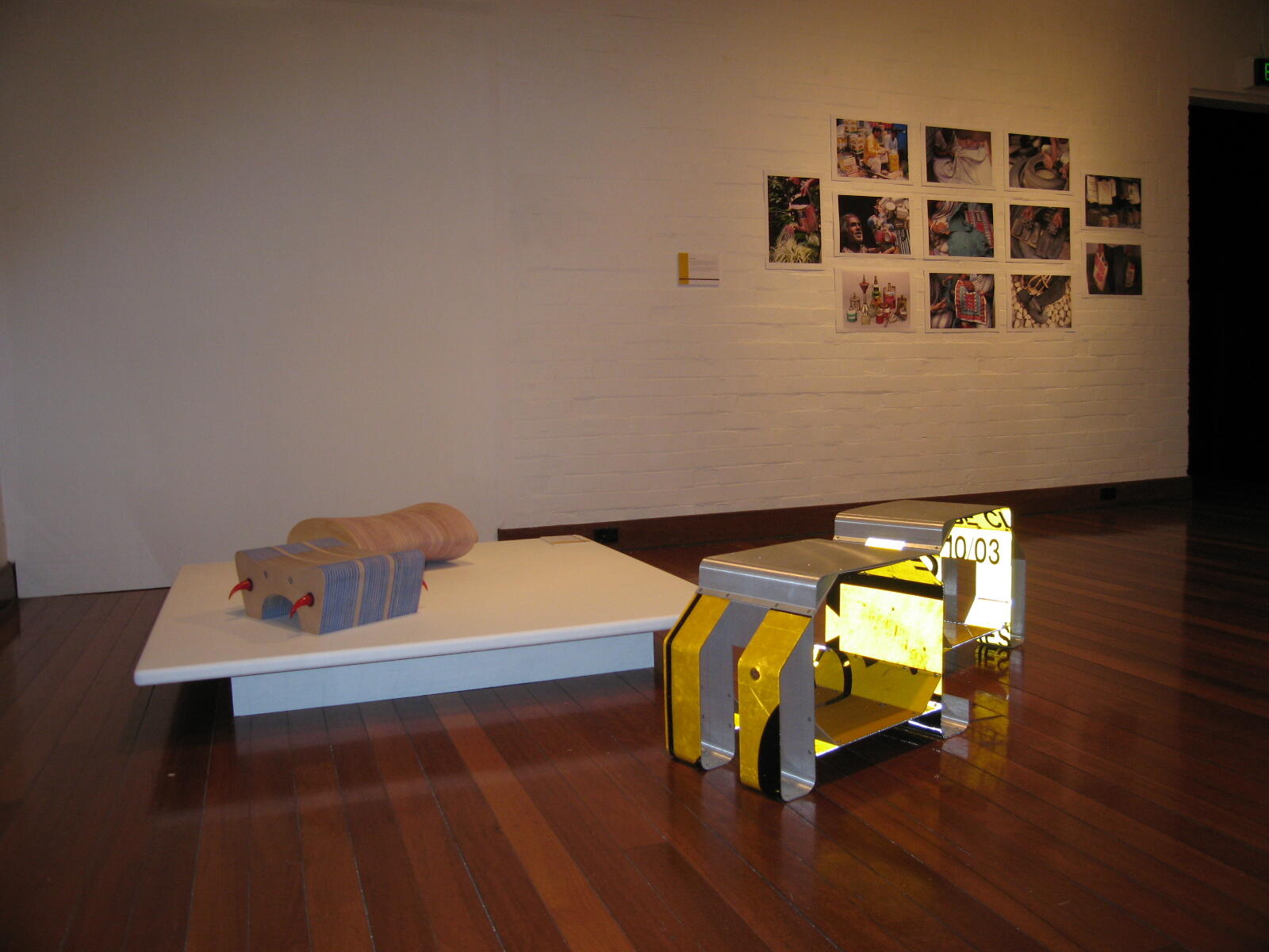 multiple artworks arranged across white gallery walls, including framed photographic prints and printed visual works installed in a structured, curated layout. With sculptural elements presented on a plinth