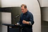 Olivier Krischer stands at a podium with a microphone, speaking passionately. The background features large, abstract geometric shapes, an artwork.