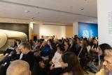 Audience sitting attentively in a gallery foyer, with a blue wall displaying "All the World's" behind them. 