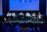 Six musicians perform on stage before a seated audience under blue lighting.