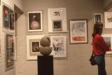 An art gallery with framed artworks displayed on white brick walls, a rounded abstract sculpture on a pedestal in the center, and a visitor viewing the exhibition.