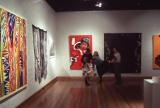 Visitors stand in an art gallery viewing colorful contemporary artworks displayed on white walls above polished wooden floors.