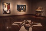 Dimly lit art gallery with polished wooden floors, two large expressive paintings mounted on brick walls, an ornate sculptural cabinet on the right, and a sprawling wing-like installation laid across the floor in the foreground.