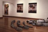 Art gallery installation with abstract paintings on a white wall, a framed figurative artwork near a striped signpost, and sculptural objects and arranged stones on a polished wooden floor.