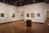 Contemporary art gallery with framed artworks displayed on white walls, a cylindrical sculpture in the center, and polished wooden floors under track lighting.