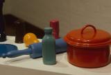 Collection of created colorful household objects, including a red lidded pot, blue rolling pin, and bottles, displayed on a white pedestal.