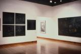 Gallery interior with large dark abstract artworks displayed on white walls and polished wooden floors.