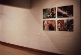 Wide view of a gallery space with a white-painted brick wall and wooden floor. Four photographic prints are mounted in a two-by-two grid on the right side of the wall, while the rest of the wall is mostly empty. The photographs depict candid outdoor scenes and close-up views of people. A small wall label is mounted beneath the display. Warm indoor lighting casts soft shadows across the wall.