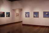 Interior of an art gallery with polished wooden floors and white brick walls displaying several framed paintings. The artworks are spaced along two intersecting walls, with soft overhead lighting illuminating the exhibition space. 