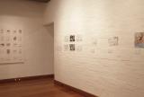 minimalist white-walled gallery interior with multiple framed artworks arranged in a horizontal sequence along the wall, featuring monochrome portrait-style prints and drawing-based works